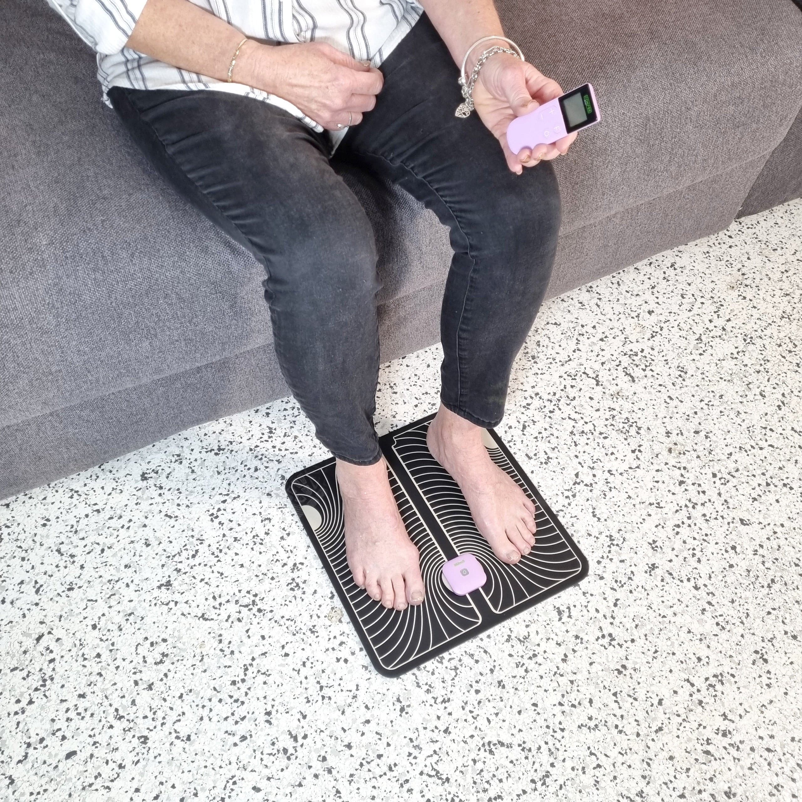 Top view of a lady sitting on a grey couch. She has her feet on a black and white foot pad that has a TENS Comfee power square attached. She is holding the TENS remote in her left hand. The TENS machine is purple in colour.
