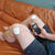 A man is sitting on an orange couch with a close up of the TENS Comfee Power3 on both is knees. The electrode pads are attached to the TENS machine through white wires. There are 2 on each knee. He is holding a Comfee Power3 in his right hand. The TENS machine is black in colour.