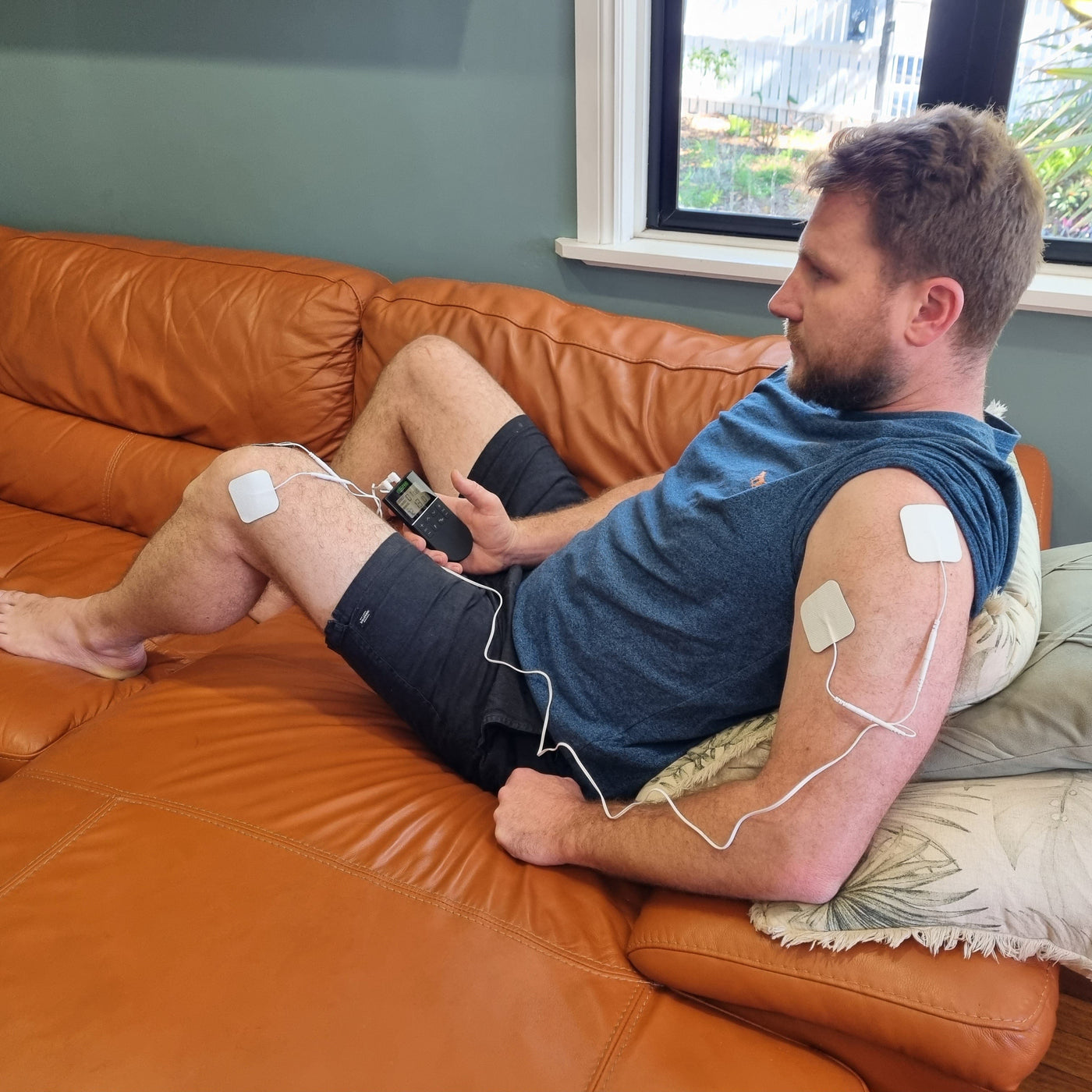 A man is sitting on an orange couch with a close up of the TENS Comfee Power3. There are 2 electrode pads on his left knee and 2 pads are on his left shoulder. The electrode pads are attached to the TENS machine through white wires. He is holding a Comfee Power3 in his right hand. The TENS machine is black in colour.