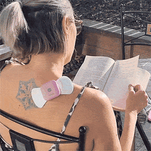 TENS Machine Comfee Power Square on a ladies shoulder. TENS Machine is pink and the lady is reading a book.