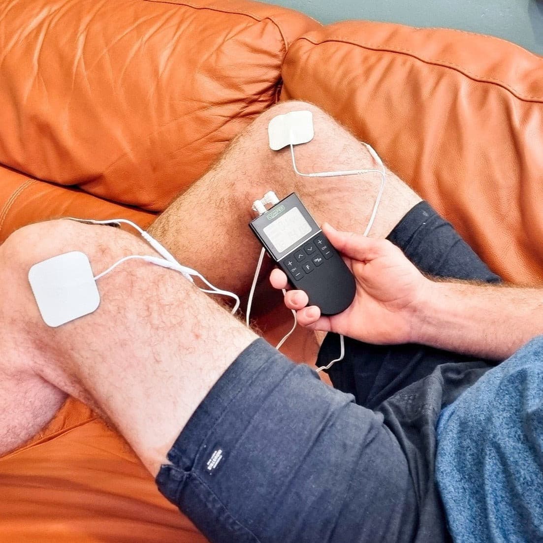 A man is sitting on an orange couch with a close up of the TENS Comfee Power3 on both is knees. The electrode pads are attached to the TENS machine through white wires. There are 2 on each knee. He is holding a Comfee Power3 in his right hand. The TENS machine is black in colour. 