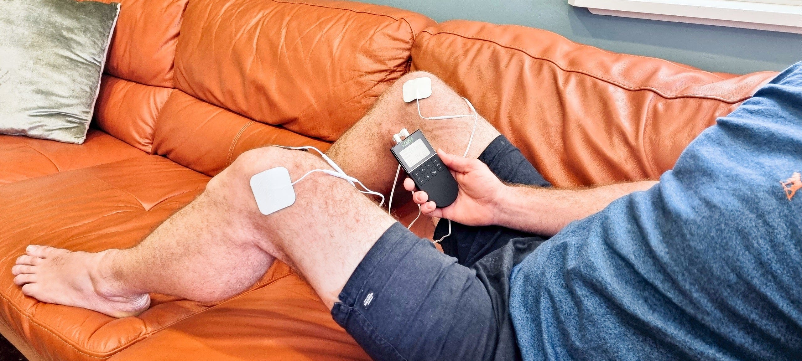 A man is sitting on an orange couch with a close up of the TENS Comfee Power3 on both is knees. The electrode pads are attached to the TENS machine through white wires. There are 2 on each knee. He is holding a Comfee Power3 in his right hand. The TENS machine is black in colour.