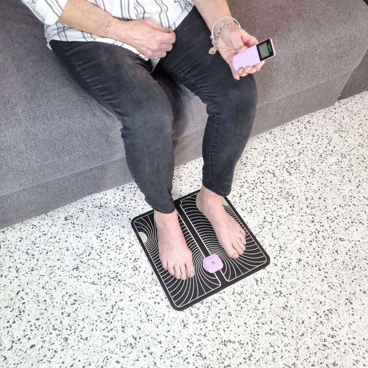 Top view of a lady sitting on a grey couch. She has her feet on a black and white foot pad that has a TENS Comfee power square attached. She is holding the TENS remote in her left hand. The TENS machine is purple in colour. 