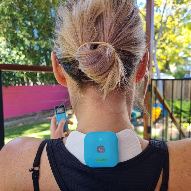 TENS Machine Comfee Power Square on the back of the neck. TENS Machine is blue and the lady is holding the remote up, directing the TENS machine from it. The TENS machine is wireless.