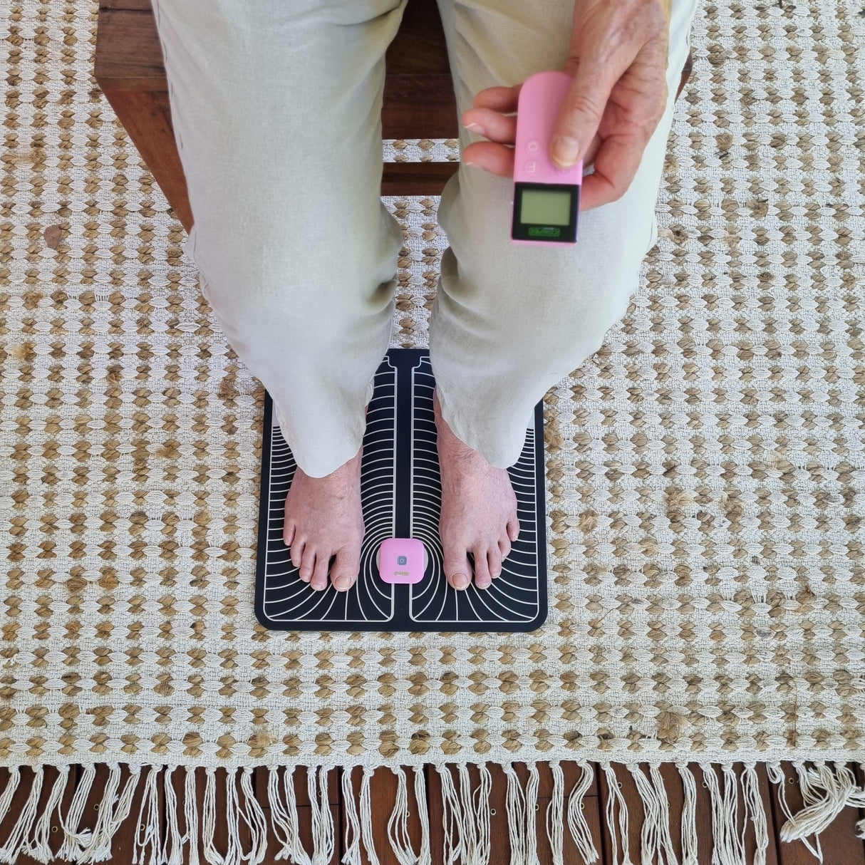 Top view of a lady sitting with her feet on a black and white footpad. The TENS Comfee power squre is clipped into the top of the foot pad. She is holding the remote in her left hand. The TENS machine and remote are pink in colour. 