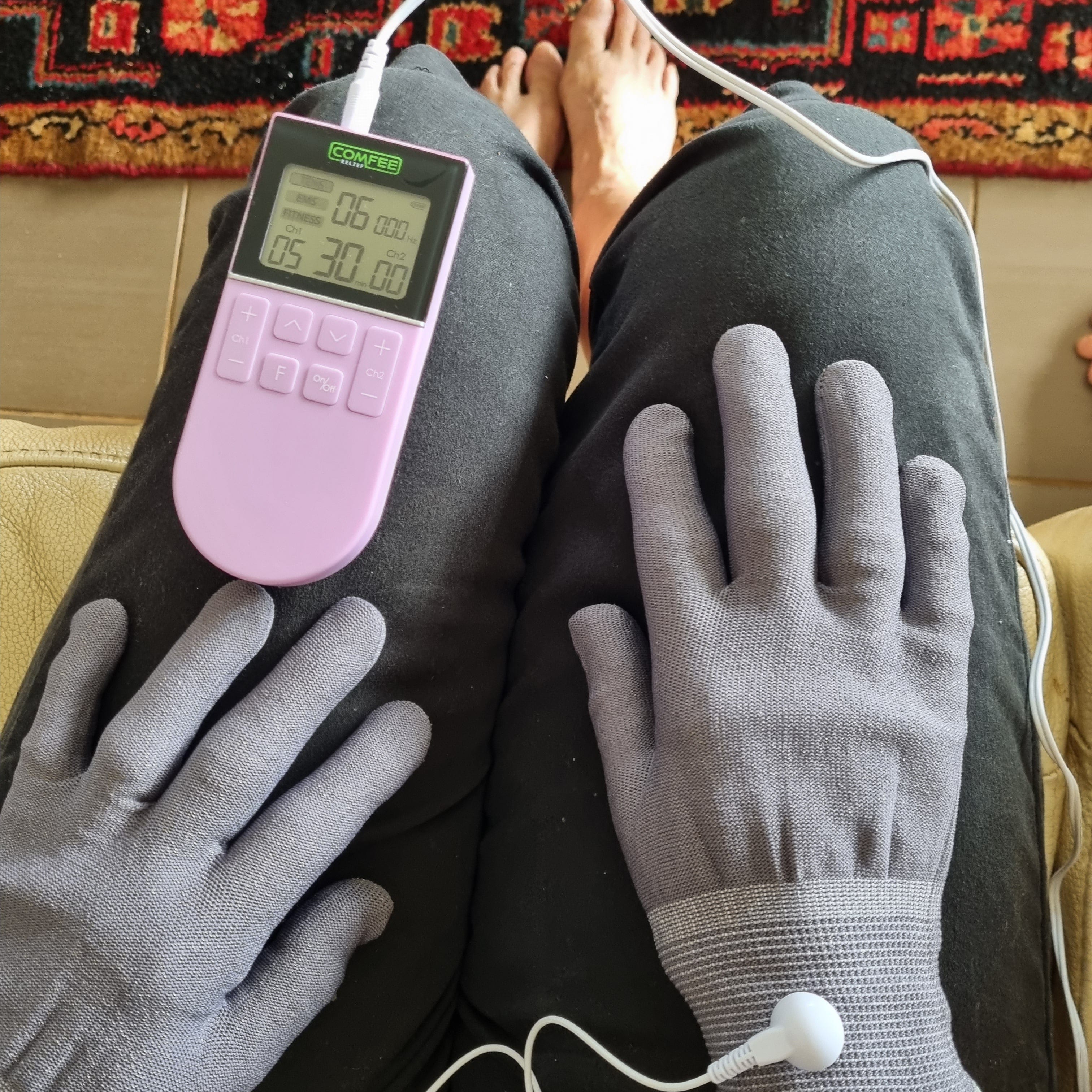 TENS Comfee power3 is sitting on the lap of a lady. There is a close up of the TENS machine along with the Pain Relief Gloves she is wearing. The Pain relief gloves are grey in colour and she is resting them on her lap. The TENS machine is on and lit up. There is a white cord attaching the gloves to the TENS machine. The TENS machine is purple in colour. 