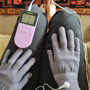 TENS Comfee power3 is sitting on the lap of a lady. There is a close up of the TENS machine along with the Pain Relief Gloves she is wearing. The Pain relief gloves are grey in colour and she is resting them on her lap. The TENS machine is on and lit up. There is a white cord attaching the gloves to the TENS machine. The TENS machine is purple in colour. 