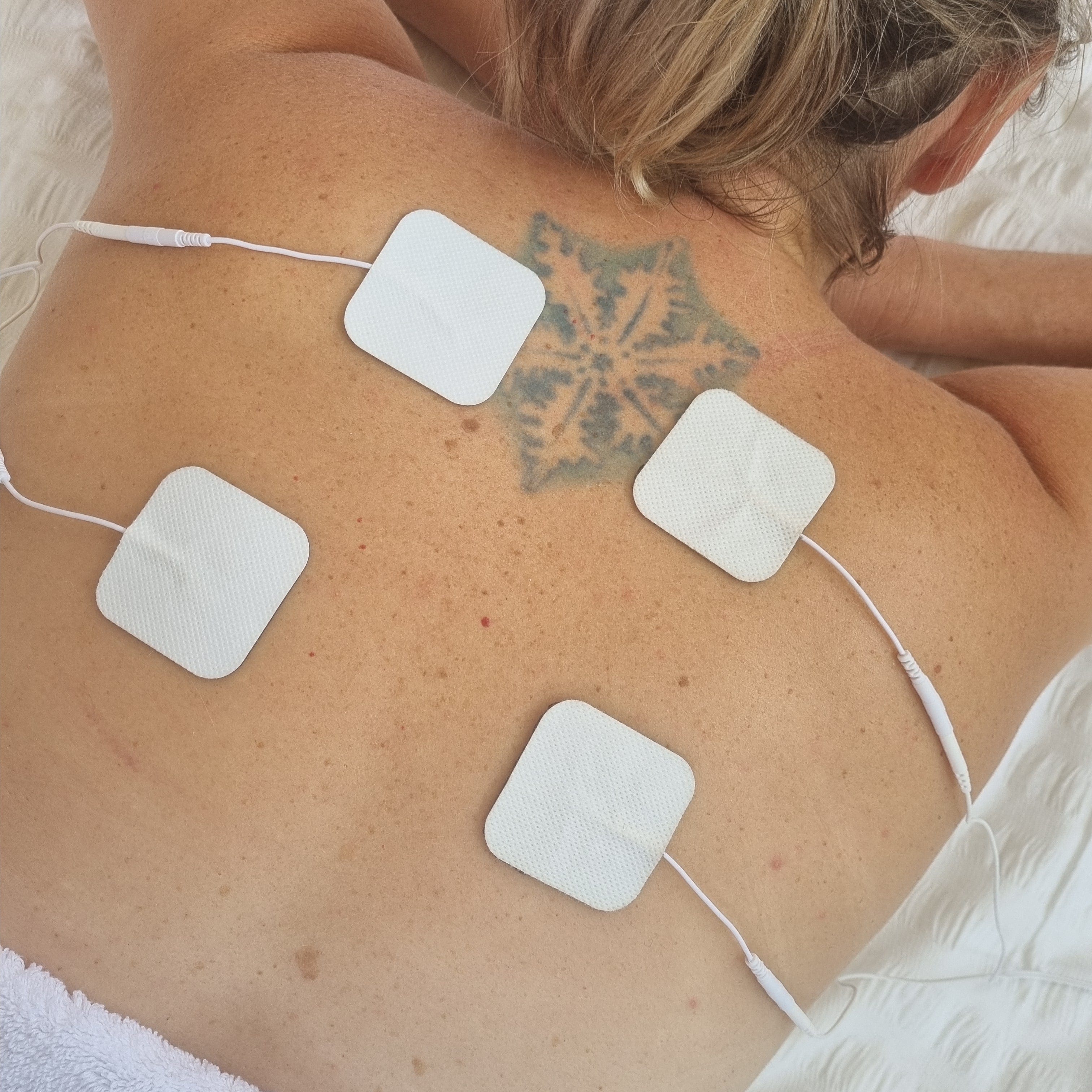 Naked back of a lady who has 4 electrode pads attached to her upper back. She is lying on a white blanket, she is blonde and fair skinned. She has a blue tattoo of a snowfake. All electrode pads are white and have white cords. 