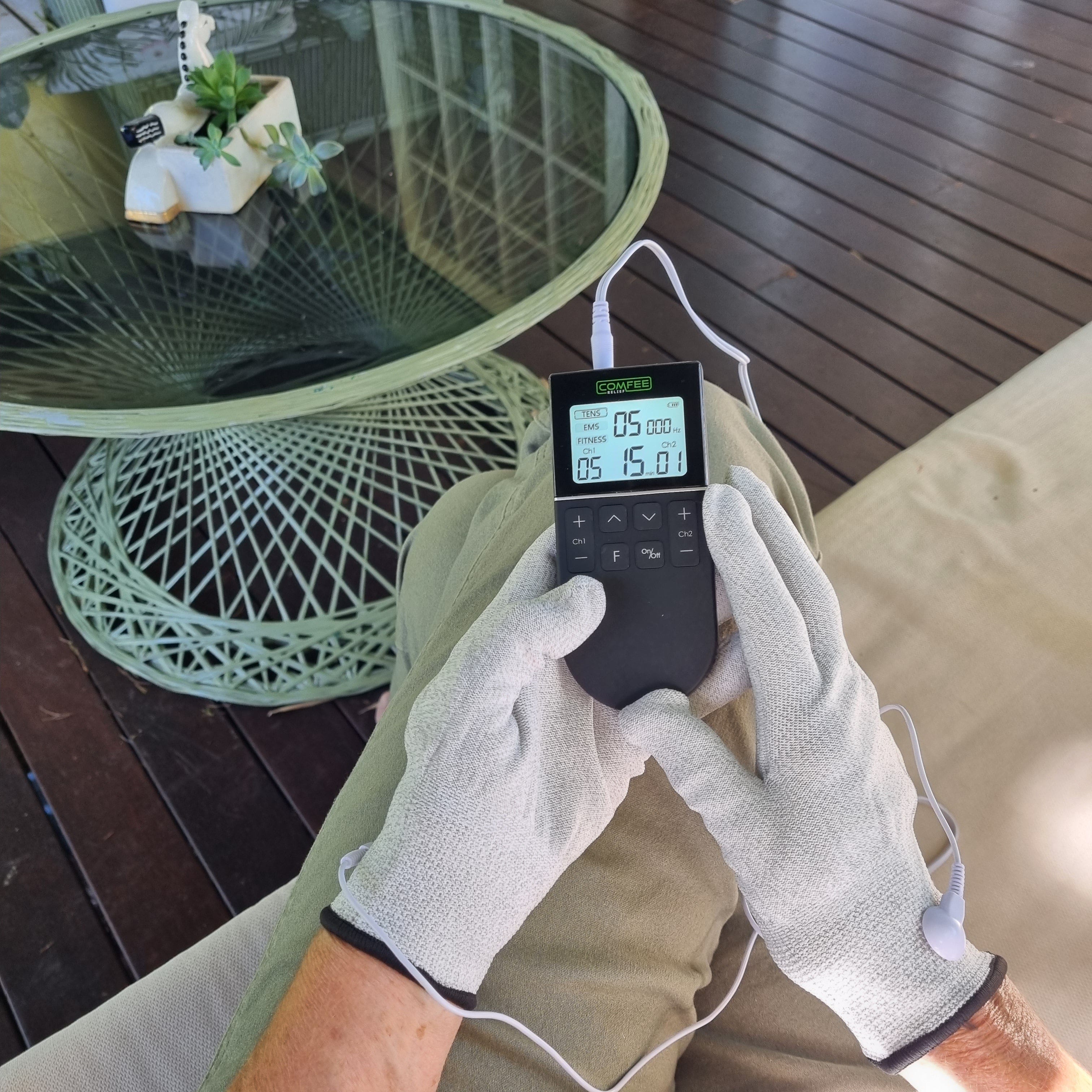 TENS Comfee power3 is being held by a lady who is also wearing the pain relief gloves. The close up only shows the lap of the lady from a top view. The Pain relief gloves are grey in colour. The TENS machine is on and lit up. There is a white cord attaching the gloves to the TENS machine. The TENS machine is black in colour. 