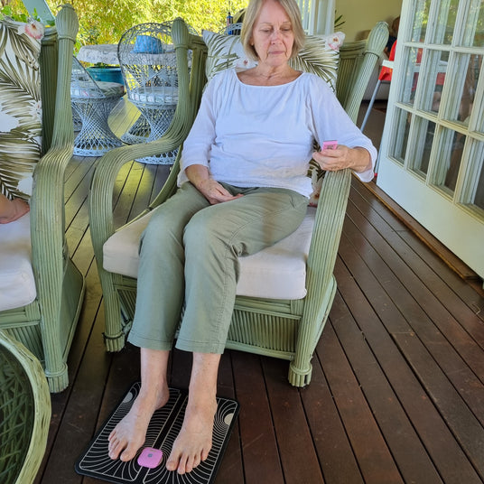 A lady sitting with her feet on a black and white footpad. The TENS Comfee power squre is clipped into the top of the foot pad. She is holding the remote in her left hand. The TENS machine and remote are pink in colour.